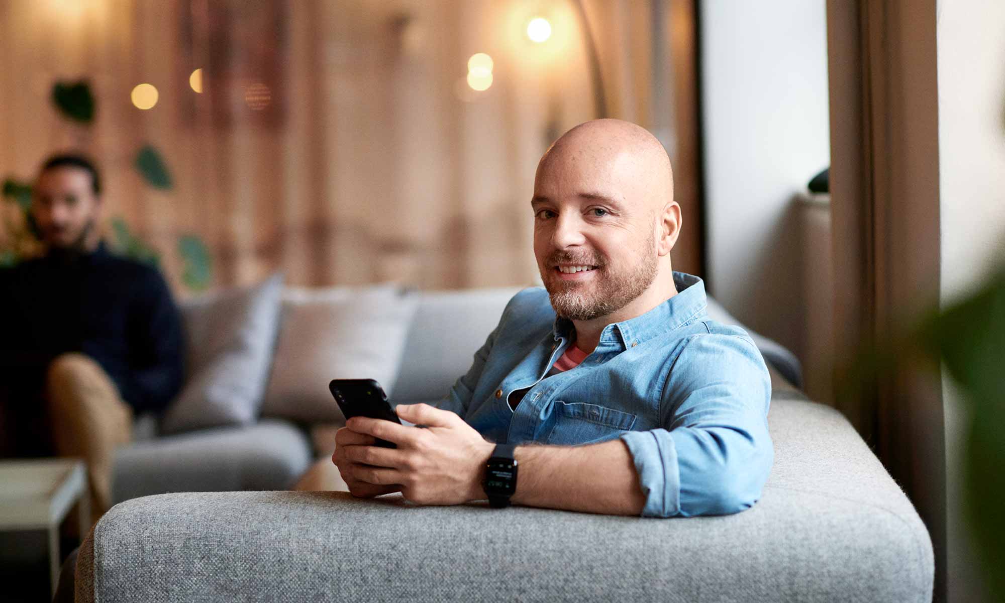 Man med blå jeansskjorta sitter i en grå soffa och håller i en mobiltelefon