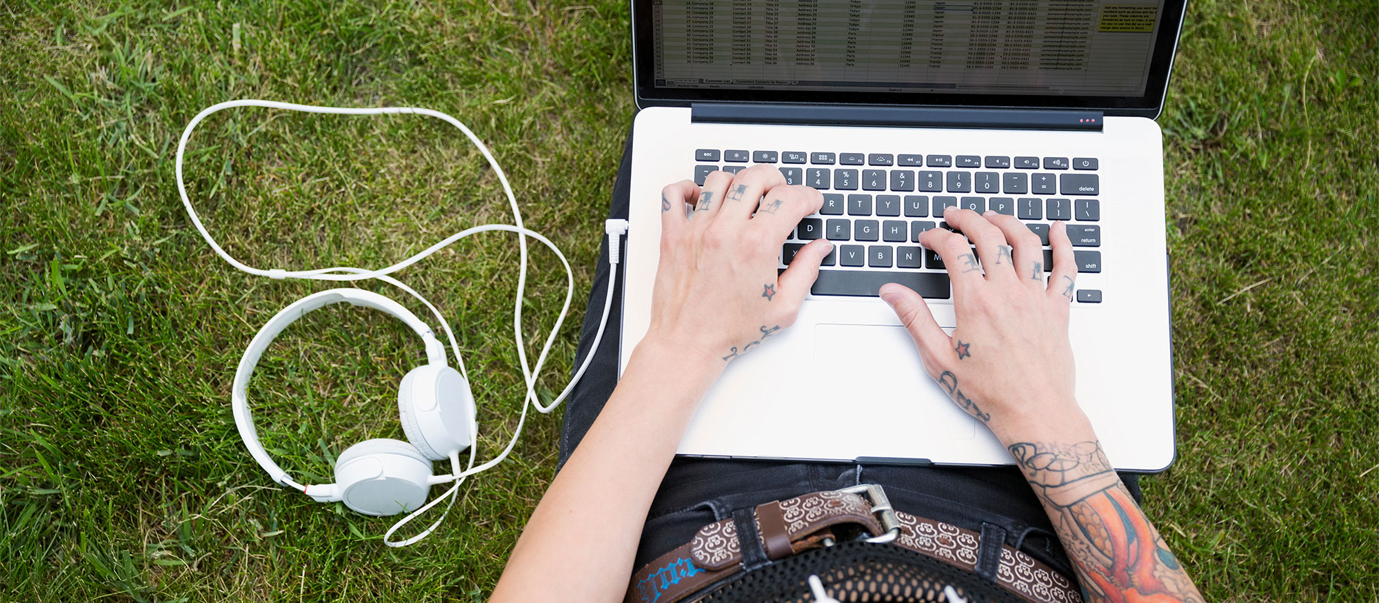 Ung person med tatueringar sitter ute i gräset med en laptop i knät